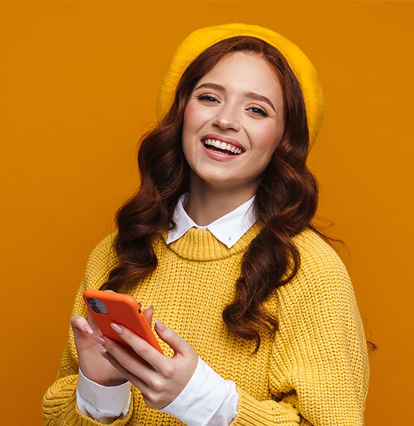 Smiling young woman with red hair wearing a yellow sweater and matching beret, holding her smartphone | Done-for-You Creative Graphics by Your Perfect Solutions